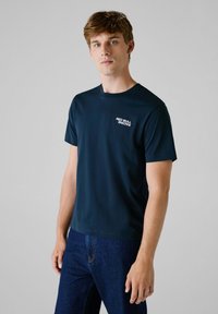 Marineblå kortermet T-skjorte laget av et glatt stoff, med en hvit "Red Bull Racing"-logo på venstre brystområde.