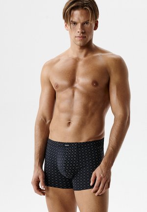 Młody muskularny mężczyzna o jasnobrązowych włosach, ubrany w czarne bokserki z białym wzorem w kratę, stojący na jasnym, jednolitym tle.