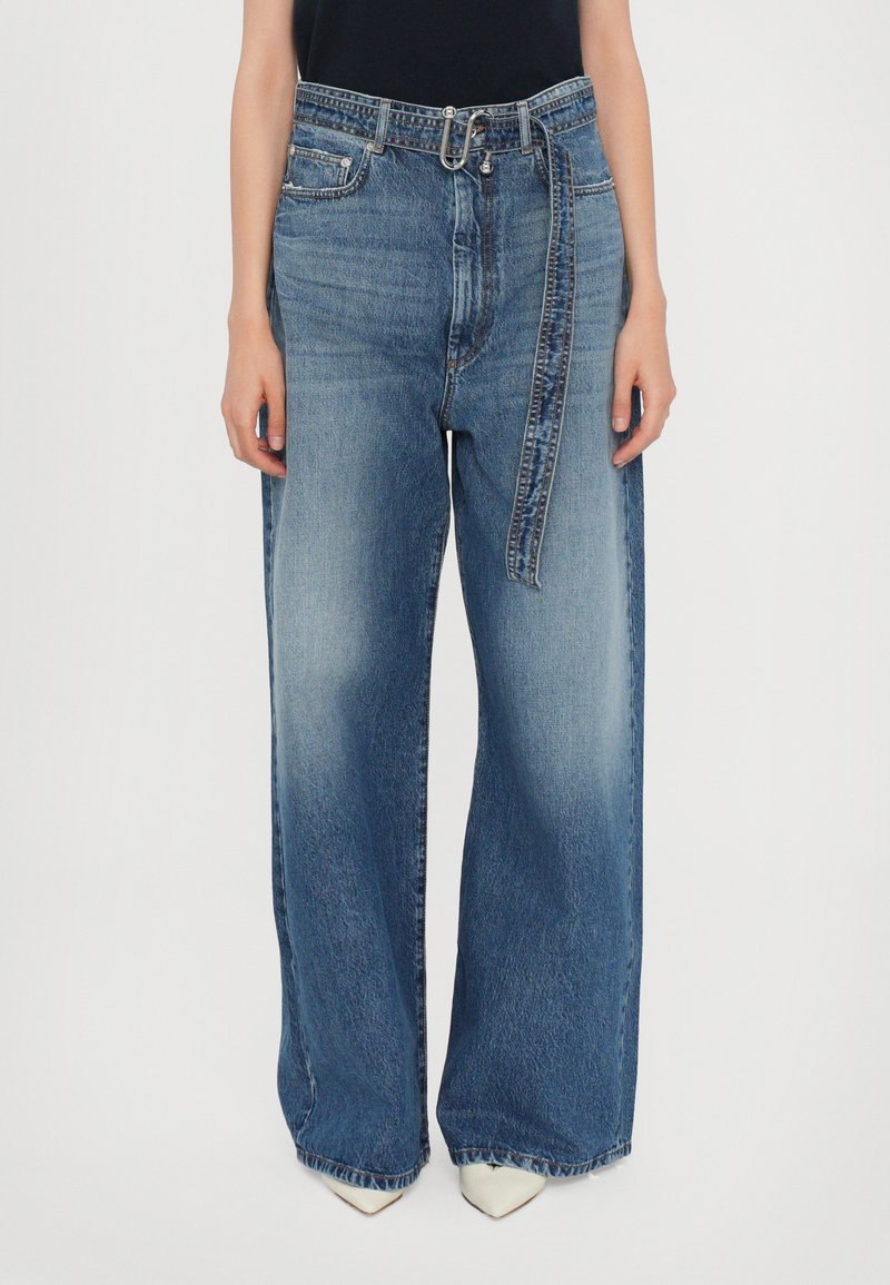 Persoon in hoge taille, wijde blauwe jeans met een denim riem en witte puntige schoenen, staand tegen een effen achtergrond.