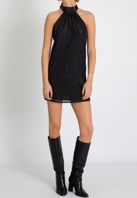 Vestido negro de halter con escote con plisados, hecho de una tela texturada brillante, combinado con botas de cuero negras hasta la rodilla.