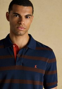 Jūras zilas polo krekls ar horizontālām brūnām svītrām. Iezīme ir sarkans apkakle, pogu plāksne un mazs izšūts logo uz krūšu daļas.