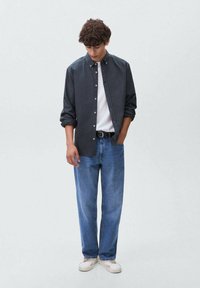 Camicia bottonata grigia scura, t-shirt bianca sotto, jeans blu e sneakers bianche. Tessuto liscio, taglio classico, leggera trama. Vestibilità casual.