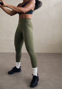 Kvinna i svart sportbh och olivgröna leggings som svingar en medicinboll i en neutral inomhusmiljö med grått golv och vägg.