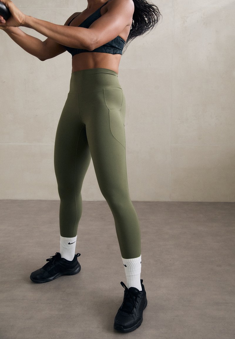Kvinna i svart sportbh och olivgröna leggings som svingar en medicinboll i en neutral inomhusmiljö med grått golv och vägg.