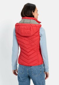 Rød quiltet vest med hætte, der har et chevronmønster og grå detaljer. Kombineret med en lyseblå langærmet bluse og denimbukser.