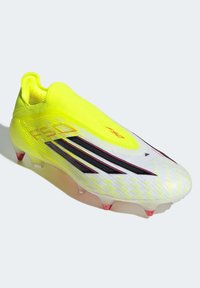 Κίτρινο και λευκό ποδοσφαιρικό παπούτσι με αεροδυναμικό σχεδιασμό, που διαθέτει μαύρες ρίγες και υφασμένη επιφάνεια για καλύτερο κράτημα. Διαφανείς τάπες με ροζ αποχρώσεις.