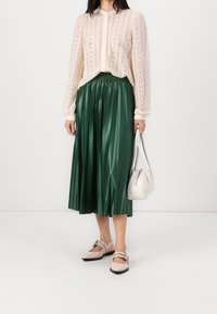 Cream lace blouse with long sleeves, green pleated midi skirt, beige shoes with straps, and a small white handbag.