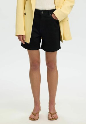 Personne portant un short en jean noir, un blazer jaune et des sandales beiges fines, debout avec une main dans la poche sur fond blanc.