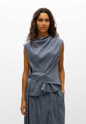 Un mannequin se tient sur un fond blanc, vêtu d’un haut gris sans manches à fines rayures, avec une taille drapée de façon asymétrique, et d’un pantalon assorti.