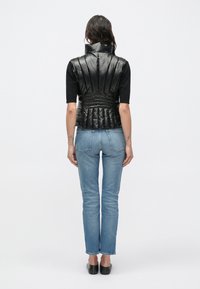 Čierna nadýchaná vesta s vysokým golierom, rebrovaným vzorovaním a zaúženým pásom, v kombinácii s modrými priliehavými džínami a čiernymi topánkami.