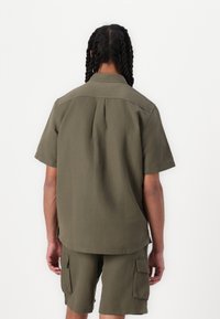Chemise verte olive à manches courtes avec un pli au dos, assortie à un short cargo. Design simple et texture tissée, sans graphiques visibles.