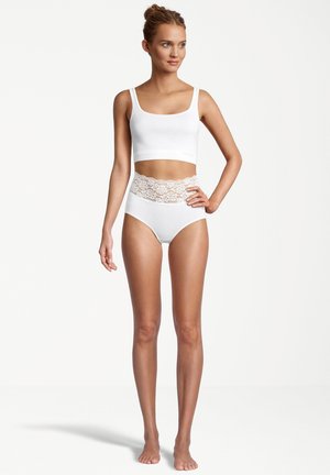 Donna in piedi a piedi nudi indossando un top corto bianco e mutandine bianche a vita alta con dettagli in pizzo su uno sfondo semplice.