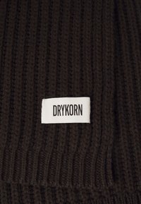Tessuto a maglia a coste di colore marrone scuro con un'etichetta rettangolare bianca cucita sopra, che mostra il nome del marchio "DRYKORN" in lettere nere.