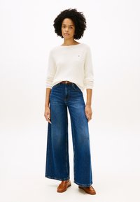 Κρεμ πουλόβερ ριπ με στρογγυλή λαιμόκοψη, συνδυασμένο με τζιν παντελόνι φαρδύ στο πόδι σε μπλε χρώμα και καφέ δερμάτινα παπούτσια.