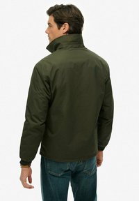 Oliwkowa kurtka z wysokim kołnierzem, elastycznymi mankietami i luźnym krojem. Wykonana z wodoodpornego materiału, charakteryzuje się gładką strukturą.