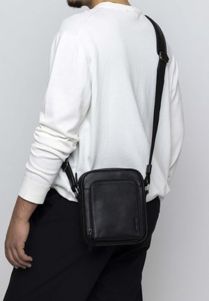 Black leather crossbody bag with a rectangular shape, zip closure, adjustable strap, and smooth texture, worn on a white long-sleeve shirt.