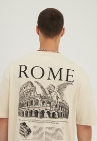 Back of a cream t-shirt with "ROME" text, detailed sketch of the Colosseum, an angel statue, and historical text about the monument.