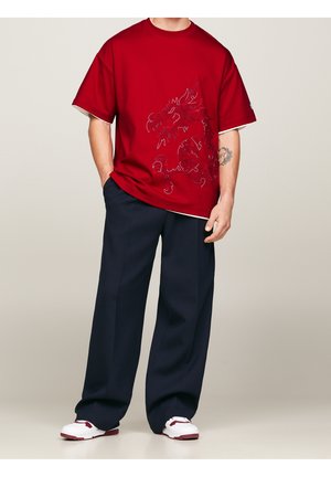 Czerwona koszulka z krótkim rękawem z haftowanym wzorem smoka, połączona z luźnymi granatowymi spodniami i białymi trampkami z czerwonymi akcentami.