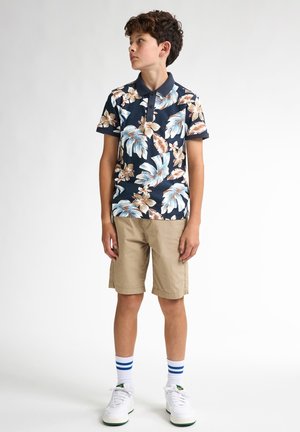 Polo de manga corta azul marino con patrón floral, pantalones cortos beige y zapatillas blancas con detalles verdes. El modelo lleva calcetas a rayas azules.