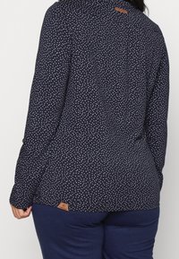 Marinblå långärmad topp med lättviktsmaterial, rundad nederkant och en liten läderlogotyp på baksidan.