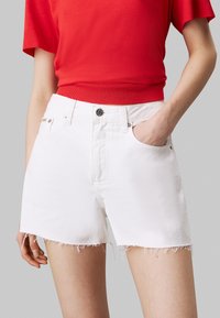Shorts de denim blanco con dobladillo deshilachado, diseño clásico de cinco bolsillos y cierre de botón metálico. Combinados con una blusa roja de manga corta.