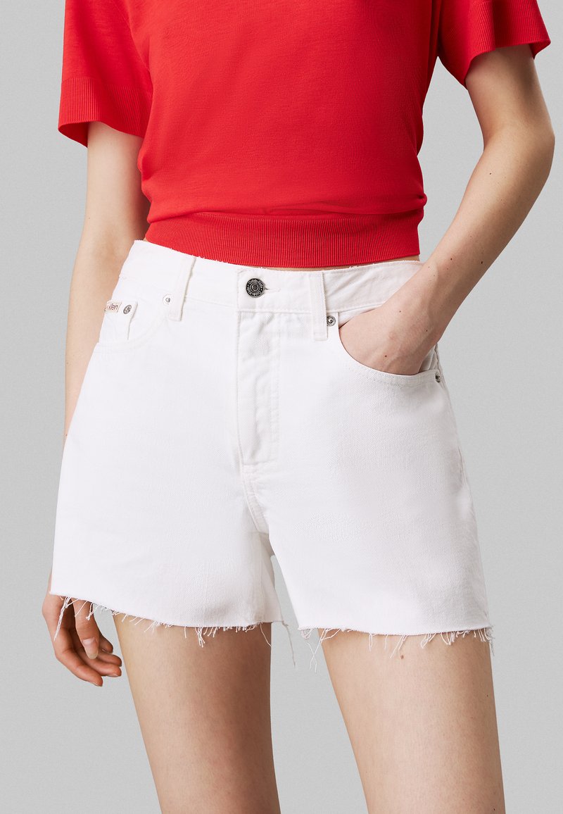 Shorts de denim blanco con dobladillo deshilachado, diseño clásico de cinco bolsillos y cierre de botón metálico. Combinados con una blusa roja de manga corta.