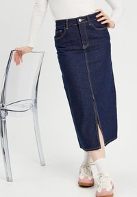 Jupe crayon en denim foncé avec fente avant, taille haute et surpiqûres contrastées. Associée à des baskets roses et blanches. Chaise transparente à côté.