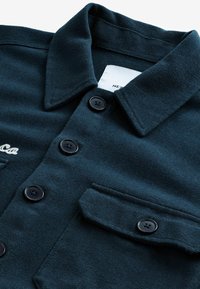 Chaqueta de canalé en verde azulado oscuro con cuello con punta, botones negros y un bolsillo en el pecho con detalle de tapa. La tela tiene un acabado texturizado.