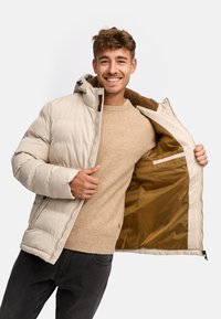 Doudoune beige avec capuche, fermeture éclair à l'avant et doublure brune texturée. Présente un extérieur lisse et une poche intérieure.
