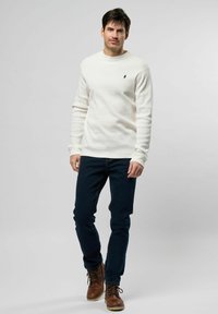 Witte geribbelde breiselsweater met ronde hals, gecombineerd met donkerblauwe slim-fit jeans en bruine veterschoenen. Subtiel zwart logo op de sweater.