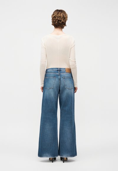 Άτομο με κοντά σγουρά μαλλιά που φοράει μπεζ μακρυμάνικο πάνω μέρος, φαρδιά μπλε τζιν και μαύρα ψηλοτάκουνα παπούτσια, στέκεται γυρισμένο με την πλάτη.