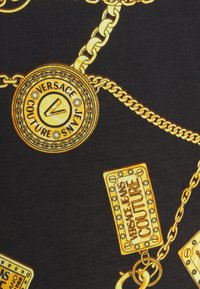 Maillons de chaîne en or avec médaillons et étiquettes Versace Jeans Couture sur fond en tissu noir.