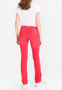 Donna che indossa pantaloni slim-fit rossi brillanti e sneakers bianche con stampe di cani, in piedi contro uno sfondo bianco uniforme, con la schiena rivolta verso lo spettatore.