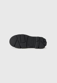Semelle de chaussure noire avec des motifs de bande de roulement géométriques et des grips texturés, présentée à plat sur un fond blanc.