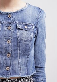 Světle modrá džínsová bunda s frayed okraji, předním zapínáním na knoflíky, kapsou s klopou, nošená přes sukně s černobílým vzorem.