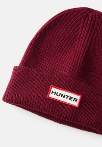 Burgundská rebrovaná pletená čiapka s prehnutým okrajom a obdĺžnikovou nášivkou loga s nápisom "HUNTER" v čiernej farbe na bielom pozadí.