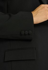 Manche de blazer noir avec trois boutons recouverts de tissu, texture lisse et détail de poche passepoilée. Le design est ajusté et structuré.