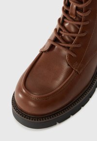 Botte en cuir marron avec bout arrondi, lacets verticaux et semelle en caoutchouc noir robuste. Détails de couture et de texture notables sur la tige.