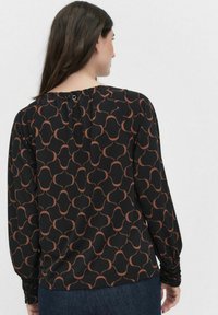 Schwarze Bluse mit geometrischem Muster in Braun, langen Ärmeln mit gerafften Manschetten und einem Schlüssel-Ausschnittdetail auf der Rückseite.