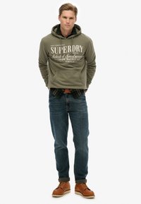 Sudadera verde con estampado blanco, superpuesta sobre una camisa de cuadros, combinada con jeans azules y botas de cuero marrón, con dobladillos enrollados.