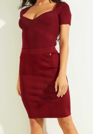 Kvinde iført en tætsiddende, ribstrikket, kortærmet bordeaux kjole med hjertesnit og knælængde nederdel mod en ensfarvet baggrund.