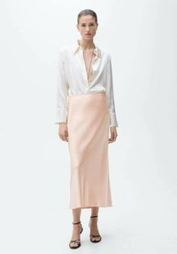 Camisă albă cu manșete lungi, combinată cu o fustă din satin roz pastel și sandale cu toc subțire negre, stând în fața unui fundal neutru.