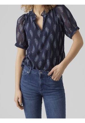 Kobieta ubrana w granatową, przejrzystą bluzkę z białymi wzorami piór i podwiniętymi krótkimi rękawami, połączoną z niebieskimi dżinsami.