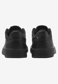 Chaussures de sport noires avec tige en cuir lisse, col rembourré et semelle en caoutchouc. Présentent un marquage minimaliste sur le talon et les côtés.