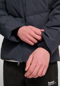 Mörkgrå puffjacka med dragkedja framtill, elastiska muddar och en slät yta, som bärs över mörka byxor. Närbild av händer som justerar ärmen.