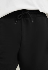 Gros plan sur un pantalon de jogging noir avec une taille élastique et un cordon noué sous une chemise blanche.