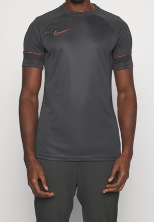 Mann trägt ein dunkelgraues kurzärmeliges Sportshirt mit orangefarbenem Nike-Logo und Besatz an den Ärmeln vor schlichtem Hintergrund.