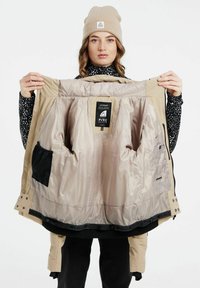 Beige isolierte Jacke mit glatter Innenauskleidung, ausgestattet mit mehreren Taschen und einem Etikett. Der Außenstoff ist leicht strukturiert.