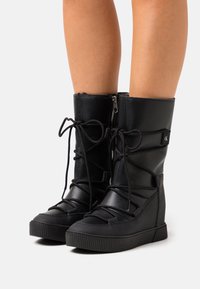 Svarta knähöga stövlar i texturerat läder med snörning och dragkedja på sidan. Har en kilklack och en gummisula med ribbad textur.