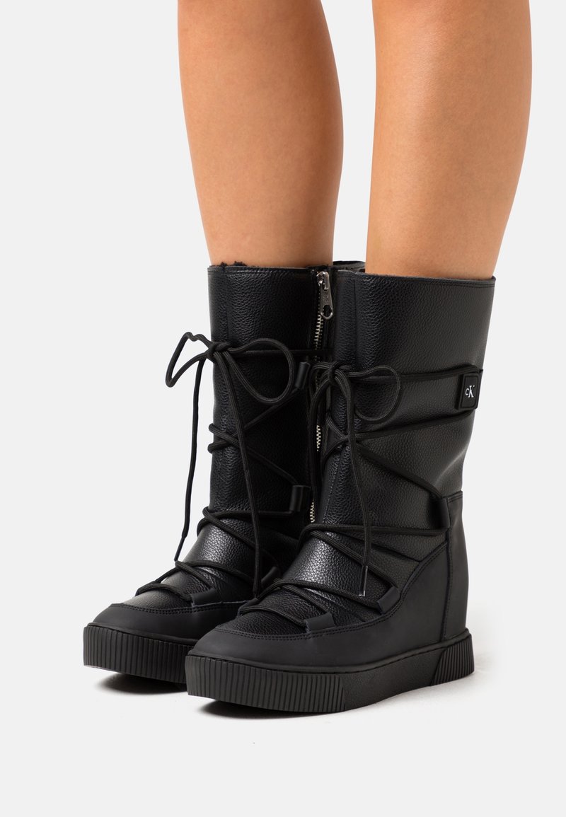 Svarta knähöga stövlar i texturerat läder med snörning och dragkedja på sidan. Har en kilklack och en gummisula med ribbad textur.